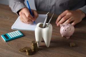 Dental tools in a tooth-shaped holder beside a piggy bank and coins, with a person writing notes, symbolizing dental care costs and financial planning for mouthguards.